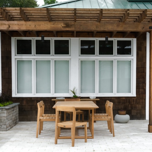 70498 Horizon Side Chair Dining Set in end view on concrete patio a teak table with 2 stacks of white plates and bowls plant against wood shingle house with large windows in background